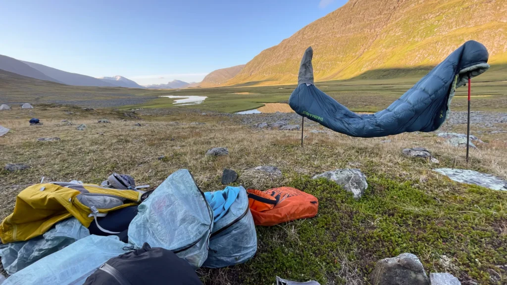 Śpiwór Therm-a-Rest Hyperion rozwieszony na kijkach trekkingowych w górskiej dolinie.